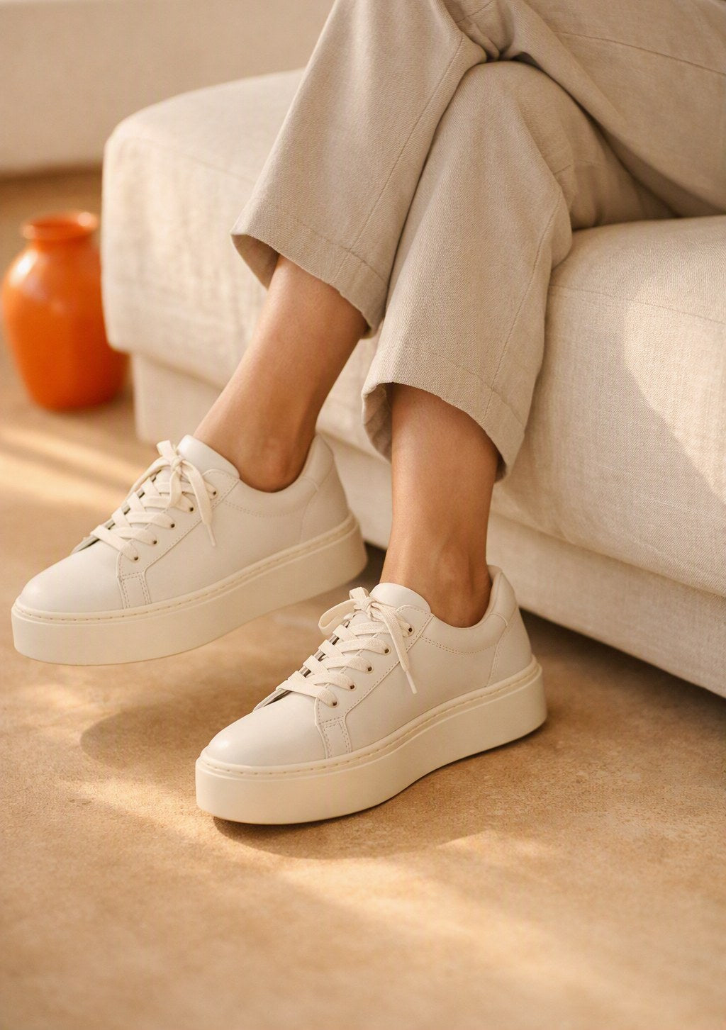 Off-white platform sneakers worn with cropped stone trousers, seated on a cream linen bench